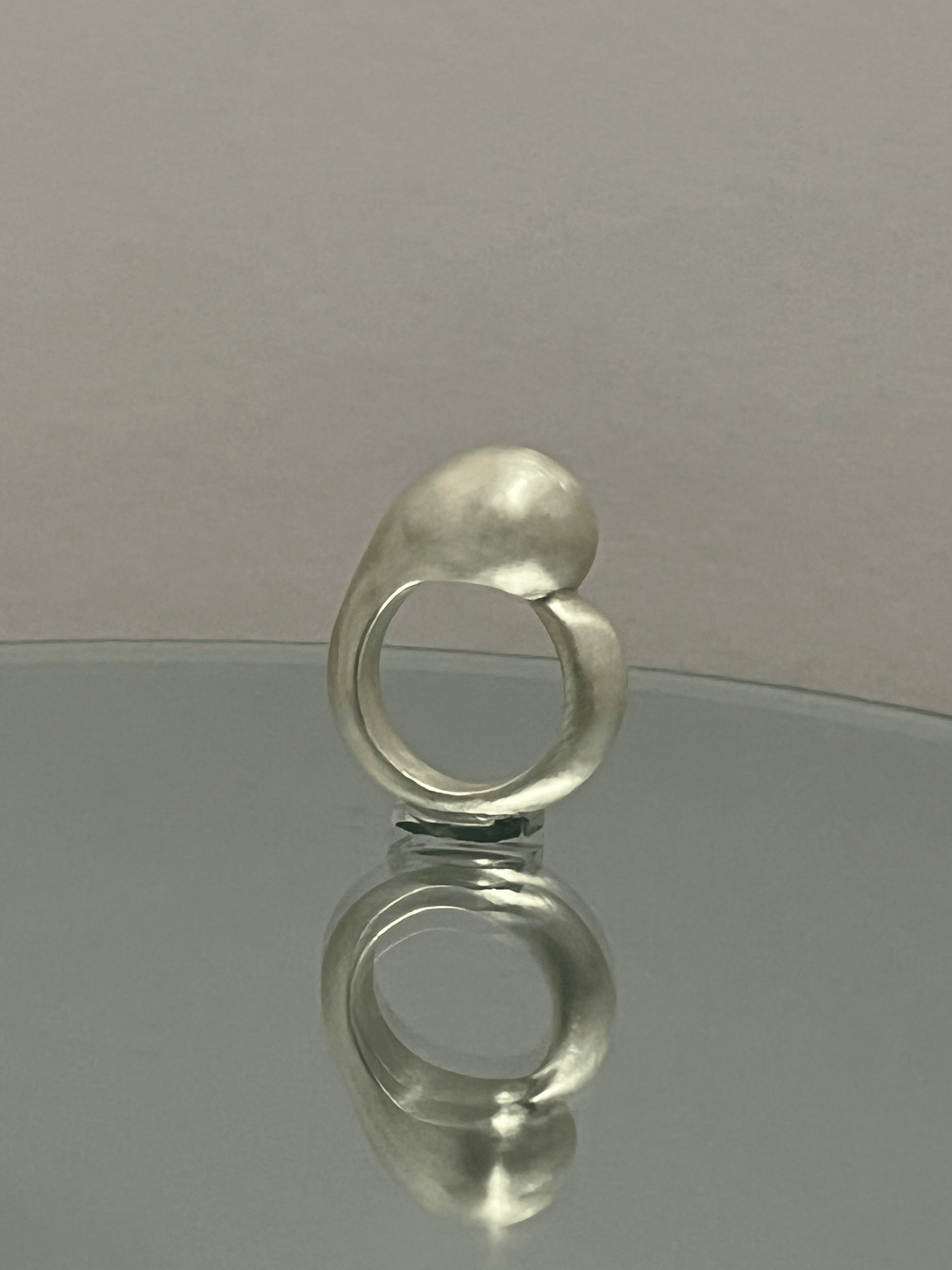 Handmade silver ring with sculptural, asymmetrical lines, placed on a mirror with a white wall backdrop. Its reflection highlights the fluid design.