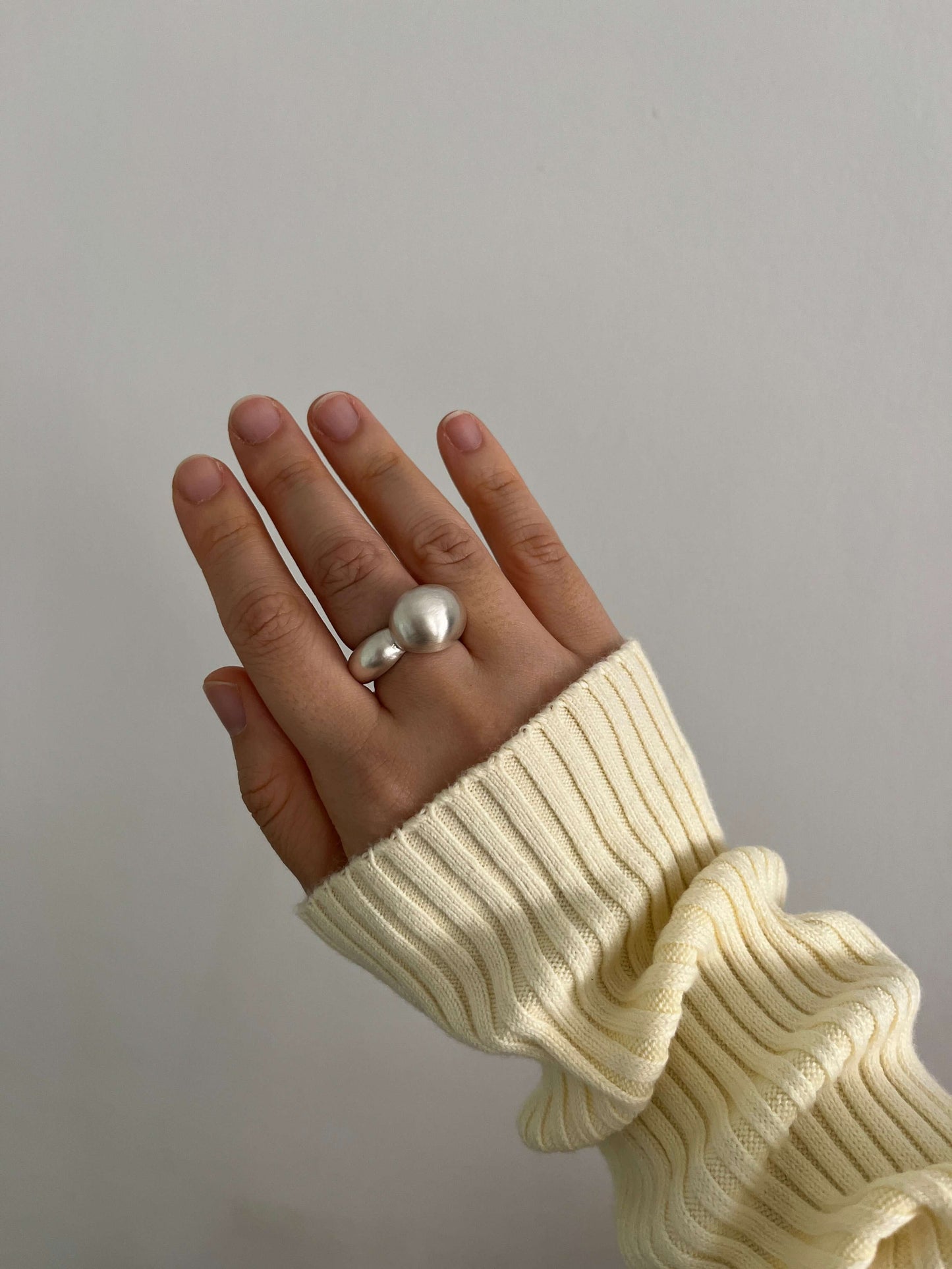 Sculptural silver ring with a bold, asymmetrical form, worn on a hand held against a white wall. A pastel yellow ribbed sleeve adds soft contrast to the ring’s polished, organic shape.