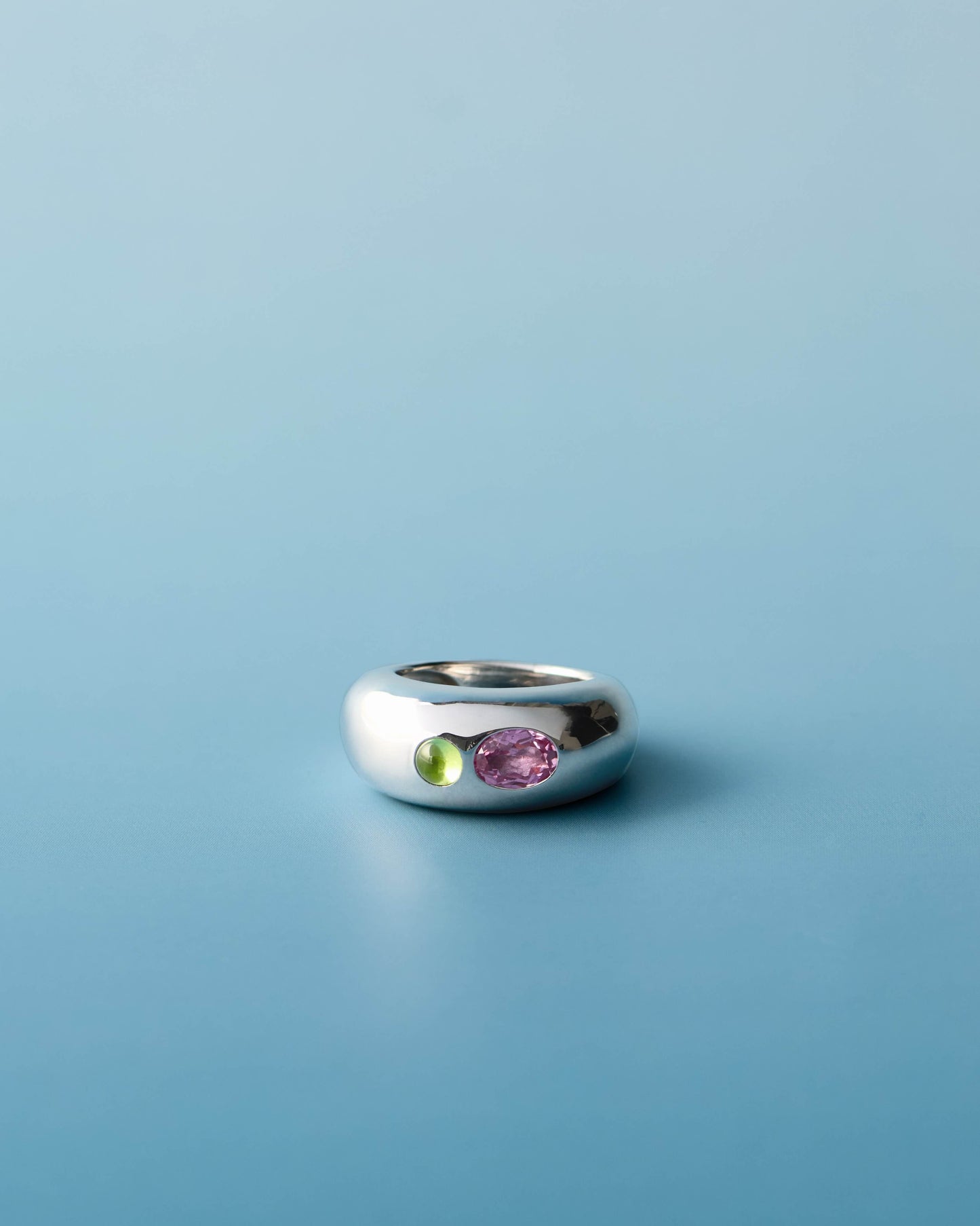 Polished silver dome ring featuring two gemstones — a natural peridot cabochon and an oval pink sapphire — set side by side. The ring is placed on a smooth, light blue background.