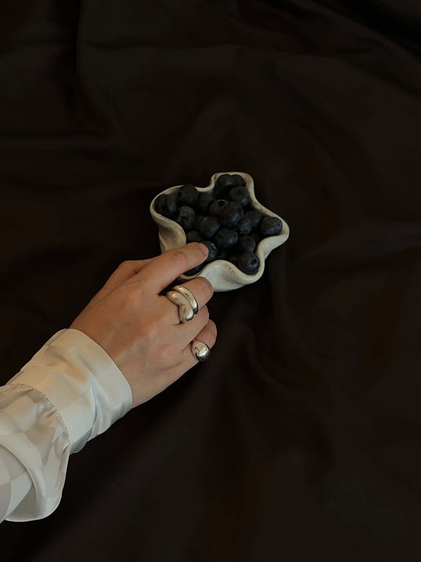 Handmade sculptural Just Be silver pinky ring with a polished dome shape, worn on a hand reaching for a star-shaped ceramic bowl filled with blueberries. The hand is adorned in a silky long sleeve, creating a beautiful contrast against the dark brown fabric background, highlighting the ring’s minimalist design and elegance.