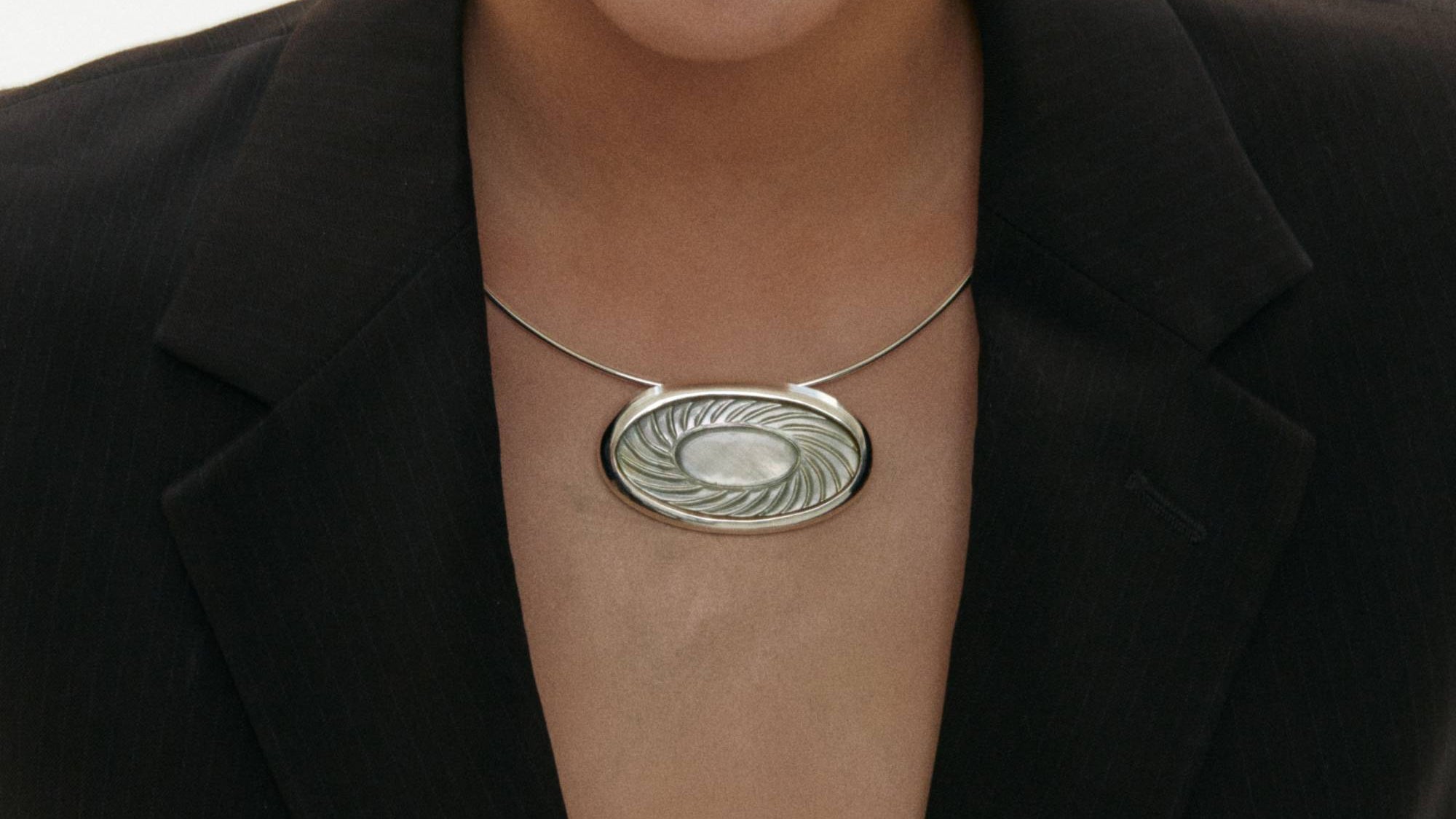 Woman wearing a brown blazer and silver torque necklace featuring a mother of shell pendant with a white background